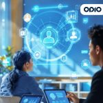 A dynamic image of three people at a desk using tablets. A man on the right and a woman in the center are focused on their screens, which display data graphs. A large, bright blue holographic interface with various icons for users, charts, and data analysis is projected over the scene.